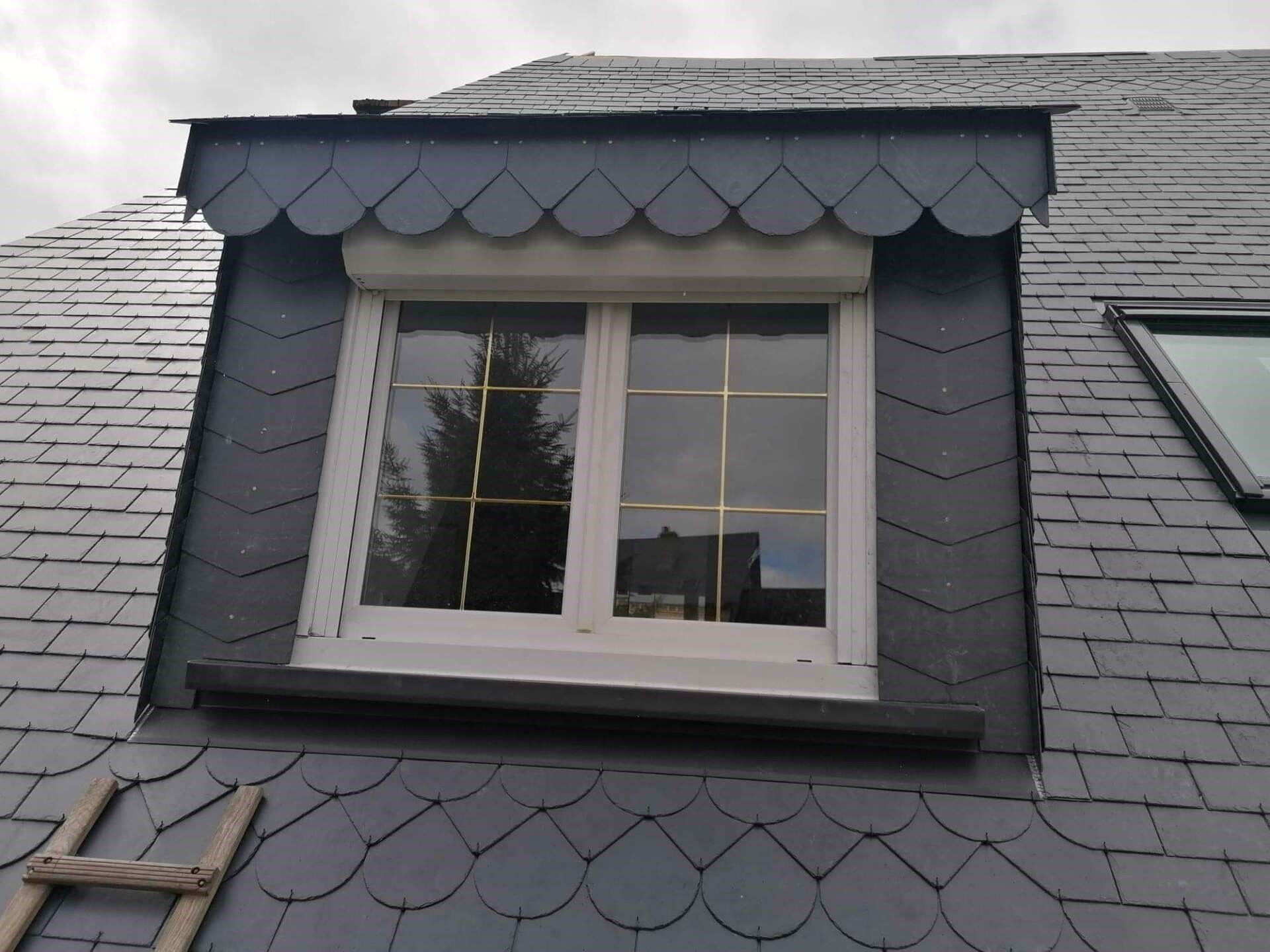 Fenêtre sur un toit en bardeaux d'ardoise grise avec moulures décoratives festonnées. Une échelle est appuyée contre le toit.
