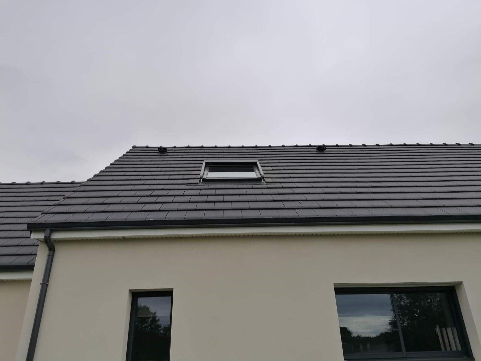 Toit en tuiles grises avec une lucarne, sur une maison beige clair sous un ciel nuageux.