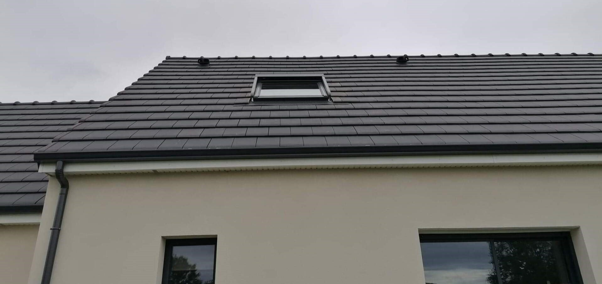 Vue extérieure d'une maison avec un toit en tuiles gris foncé, un puits de lumière et des murs de couleur claire.