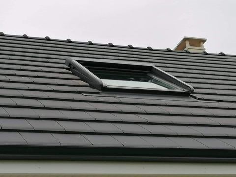 Puits de lumière sur un toit en tuiles gris foncé avec une cheminée et un ciel couvert.