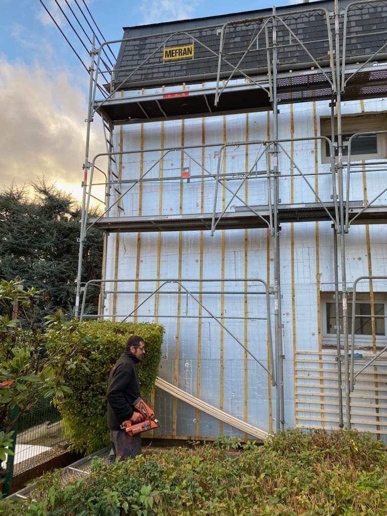 Un homme travaille sur l'extérieur d'une maison avec un échafaudage, isolation visible.