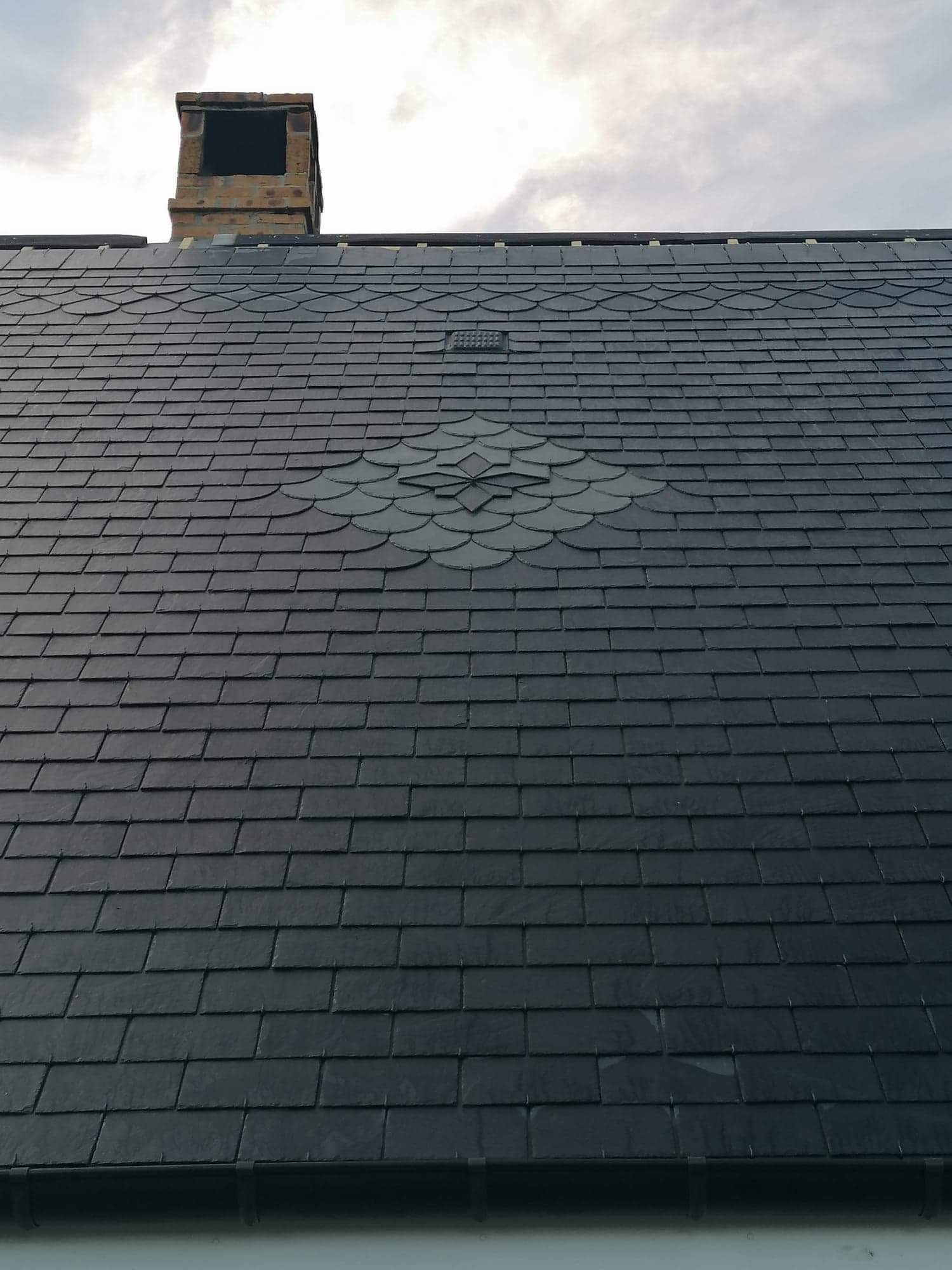 Toiture en ardoise foncée avec une zone endommagée, cheminée en arrière-plan, ciel nuageux.