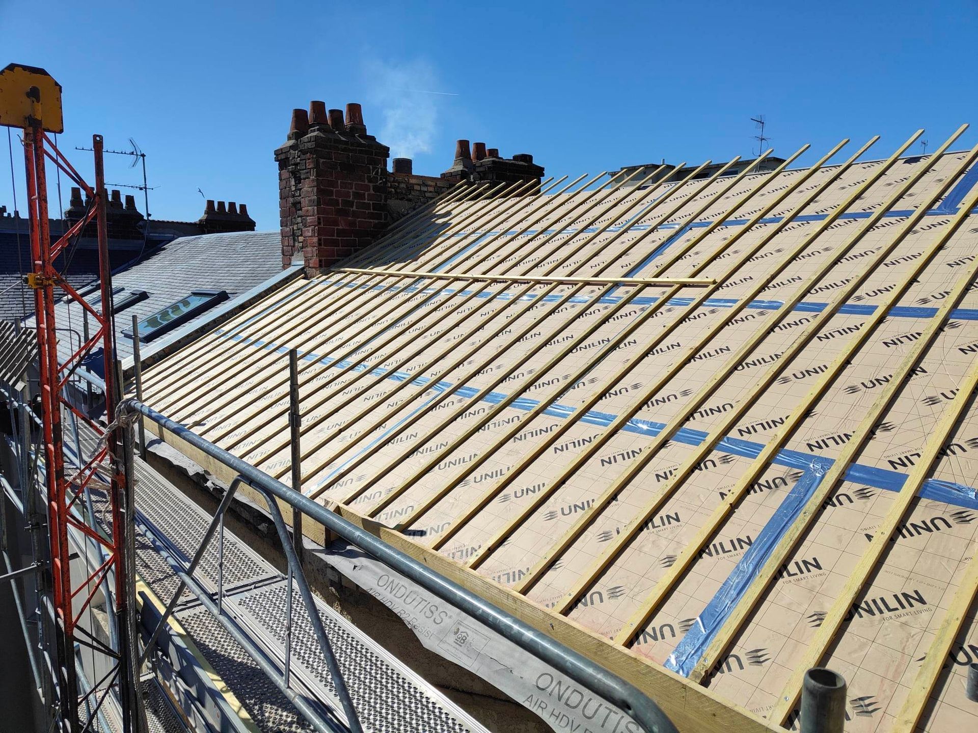 Toit en construction avec de nouvelles lattes de bois, des échafaudages et un ciel bleu clair.