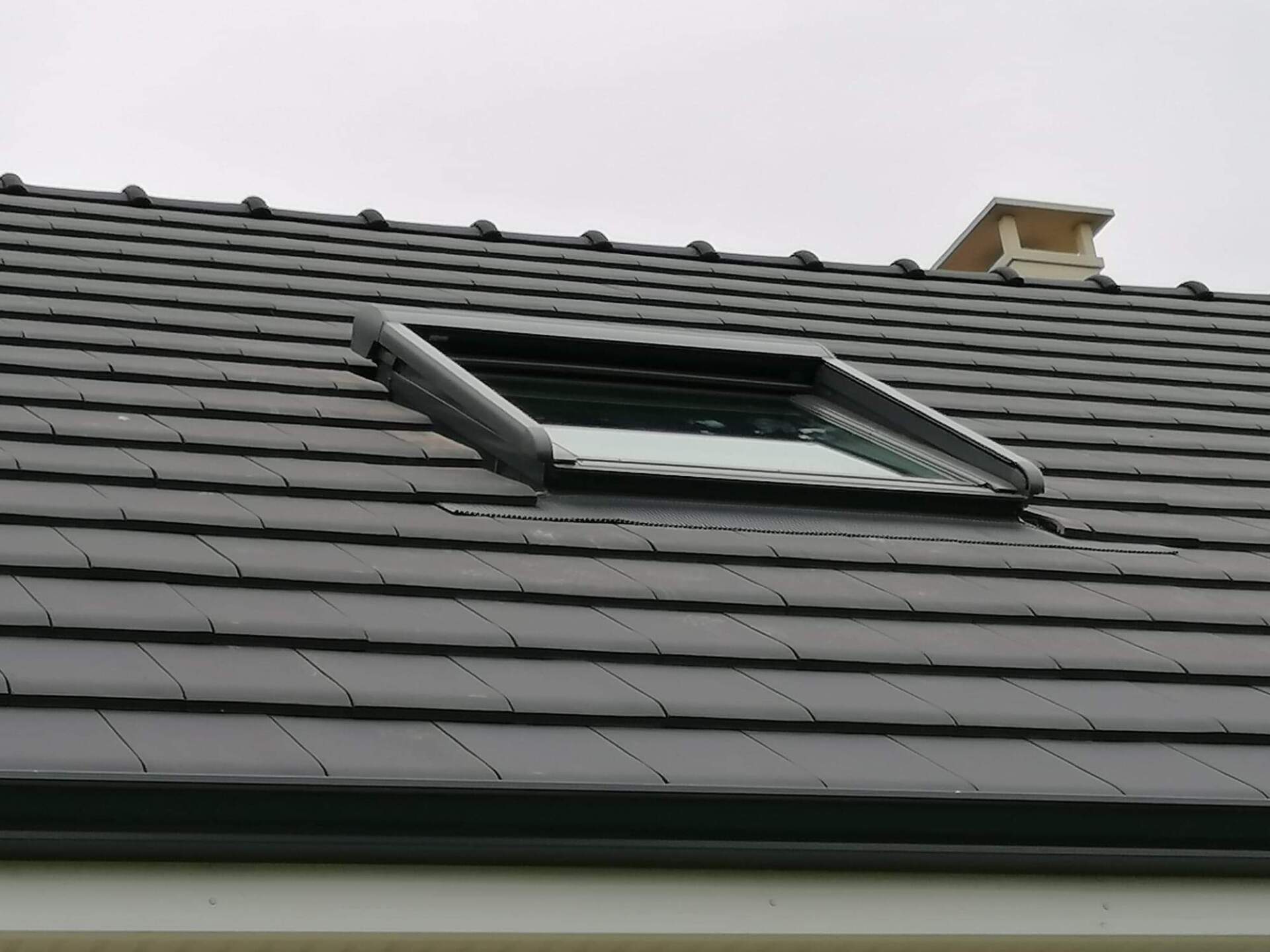 Puits de lumière sur un toit en tuiles sombres. Une cheminée blanche apparaît en arrière-plan, sous un ciel gris.