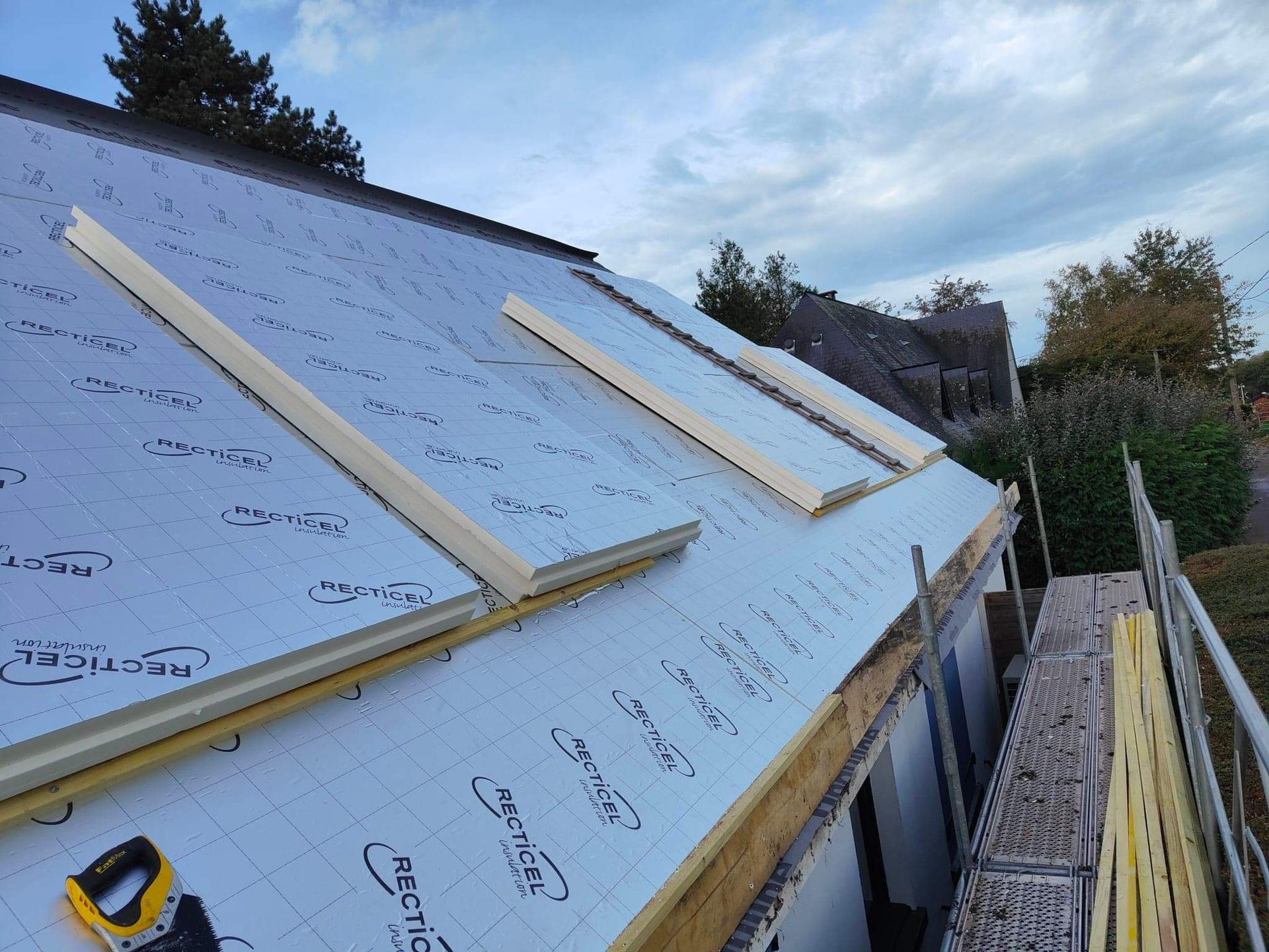 Toiture en construction avec isolation, poutres en bois et scie à main. Extérieur sous un ciel nuageux.