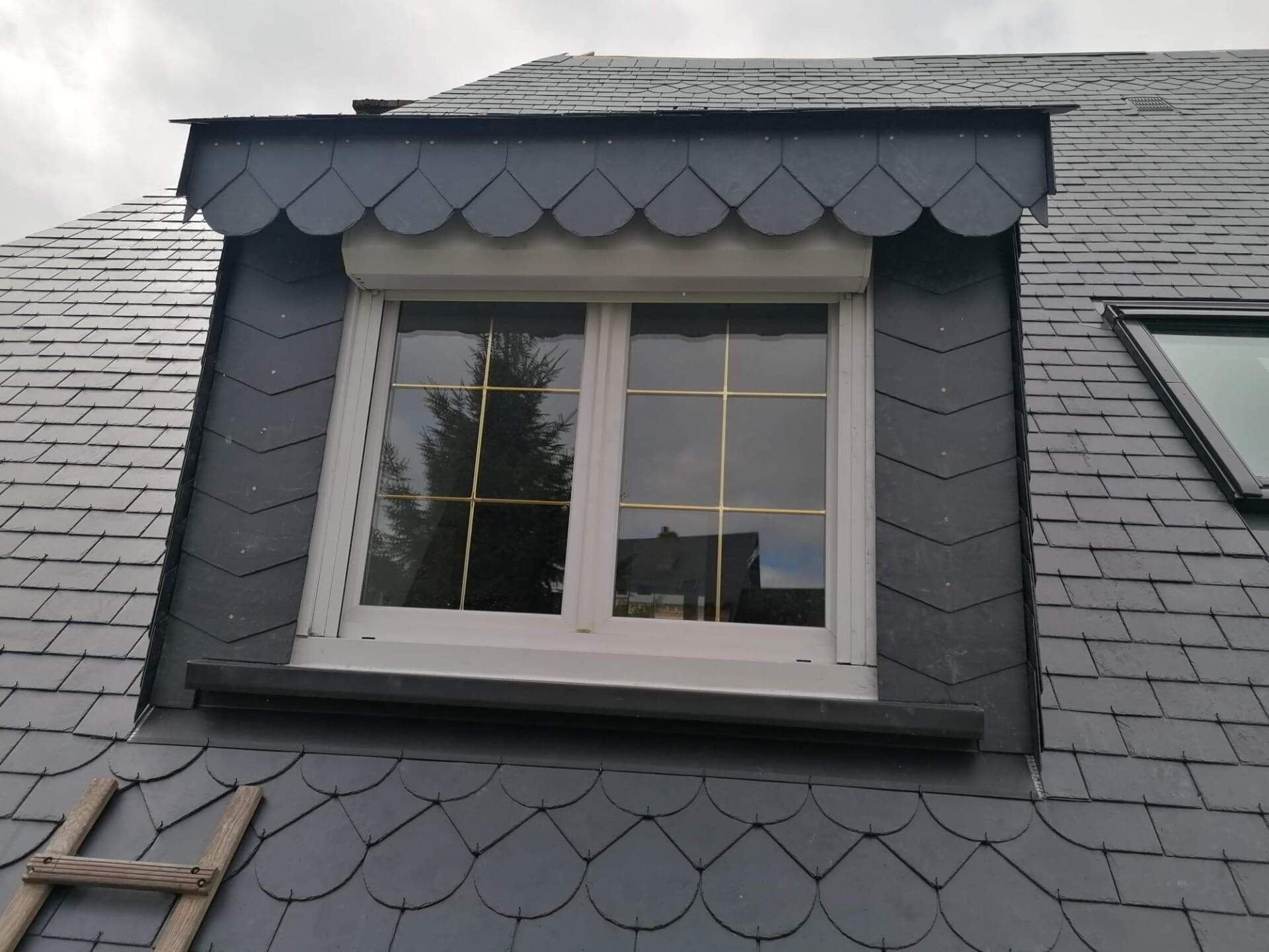 Lucarne sur toit en ardoise, cadre blanc, à bord festonné, et échelle partielle.