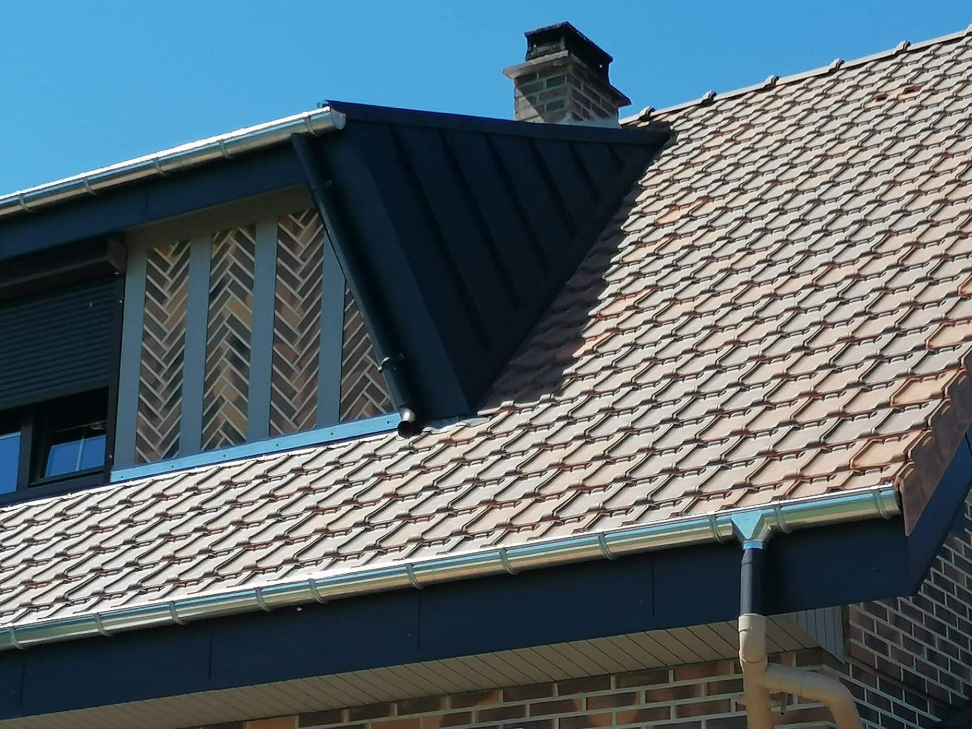 Maison en brique avec toit en tuiles brunes, section de toit en métal noir, motif à chevrons en brique, ciel bleu.