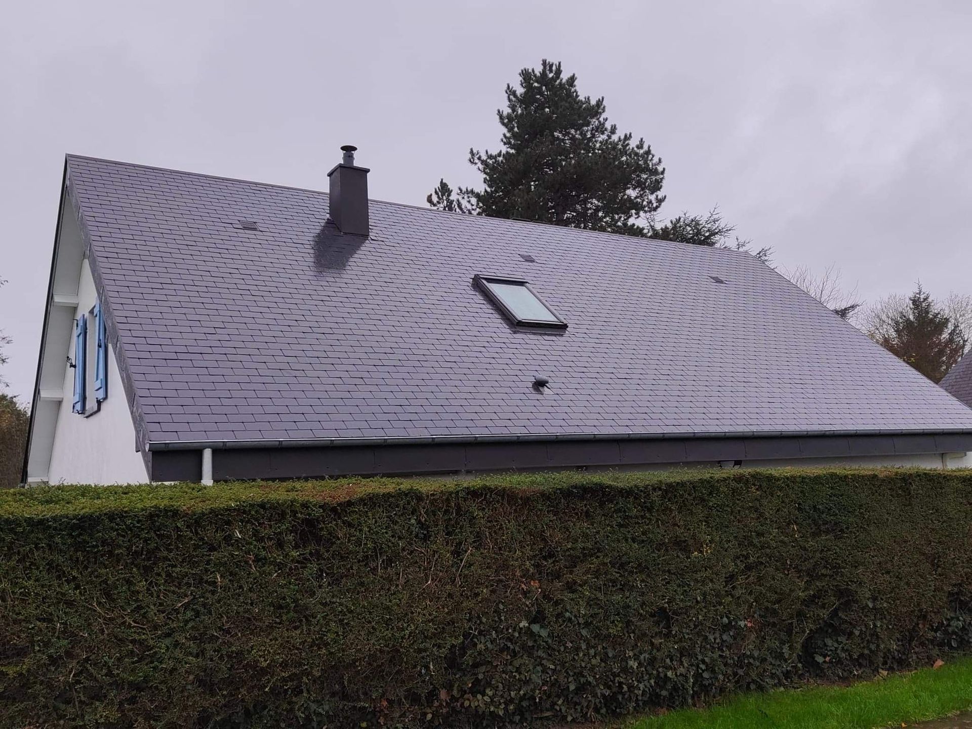 Une maison avec un toit sombre à motifs, une lucarne et une cheminée, derrière une haie verte sous un ciel couvert.
