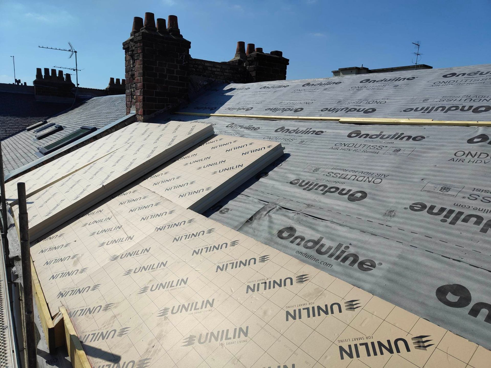 Toiture en construction ; sous-couche noire et beige visible, à côté d'une cheminée. Journée ensoleillée.