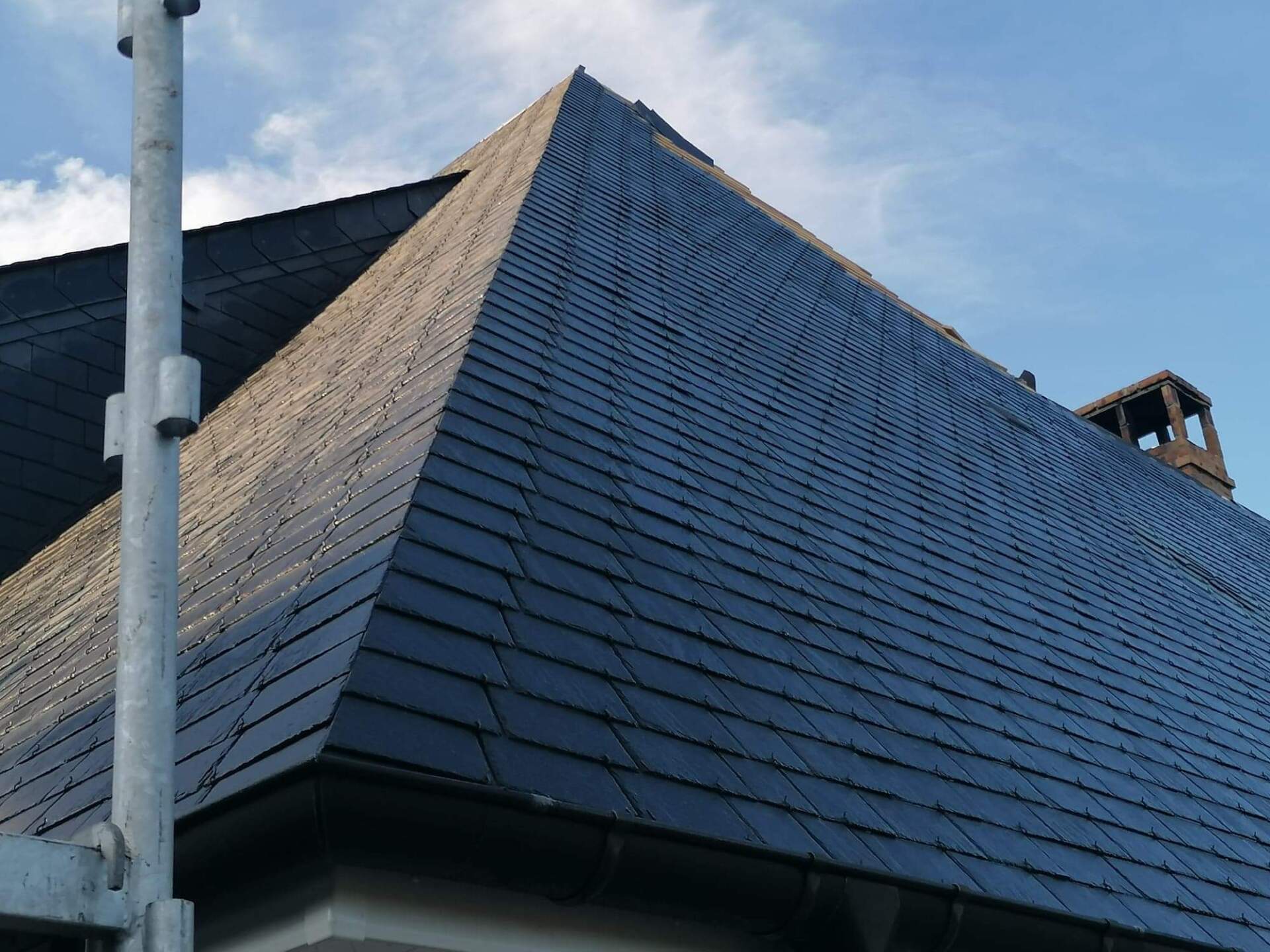 Toit en tuiles gris foncé avec cheminée et ciel bleu. Échafaudage partiellement visible.