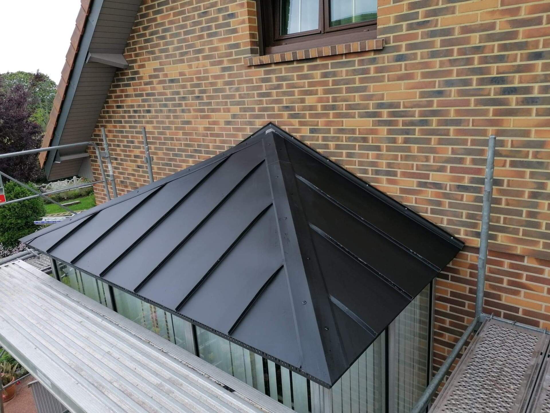 Toiture métallique noire sur une structure en forme de hangar à côté d'un bâtiment en briques. Échafaudage visible.