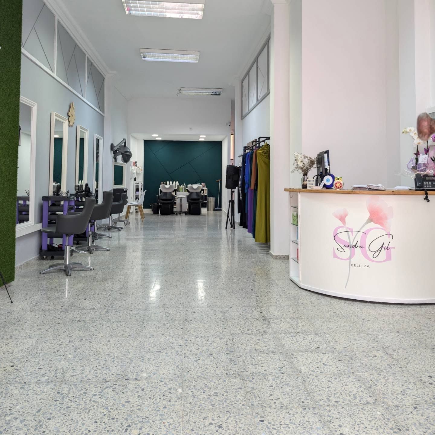 Interior de peluquería con estaciones de peinado, área de espera con escritorio y exhibición de ropa colorida.