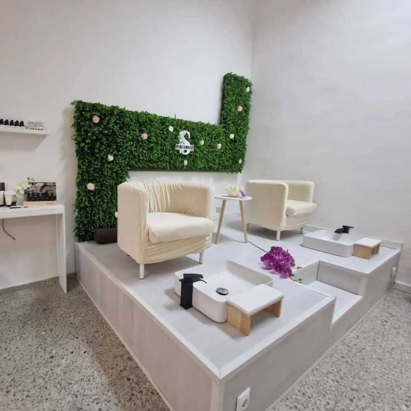Interior de salón de uñas con asientos color crema sobre una plataforma blanca. Pared con detalles verdes y adornos florales.