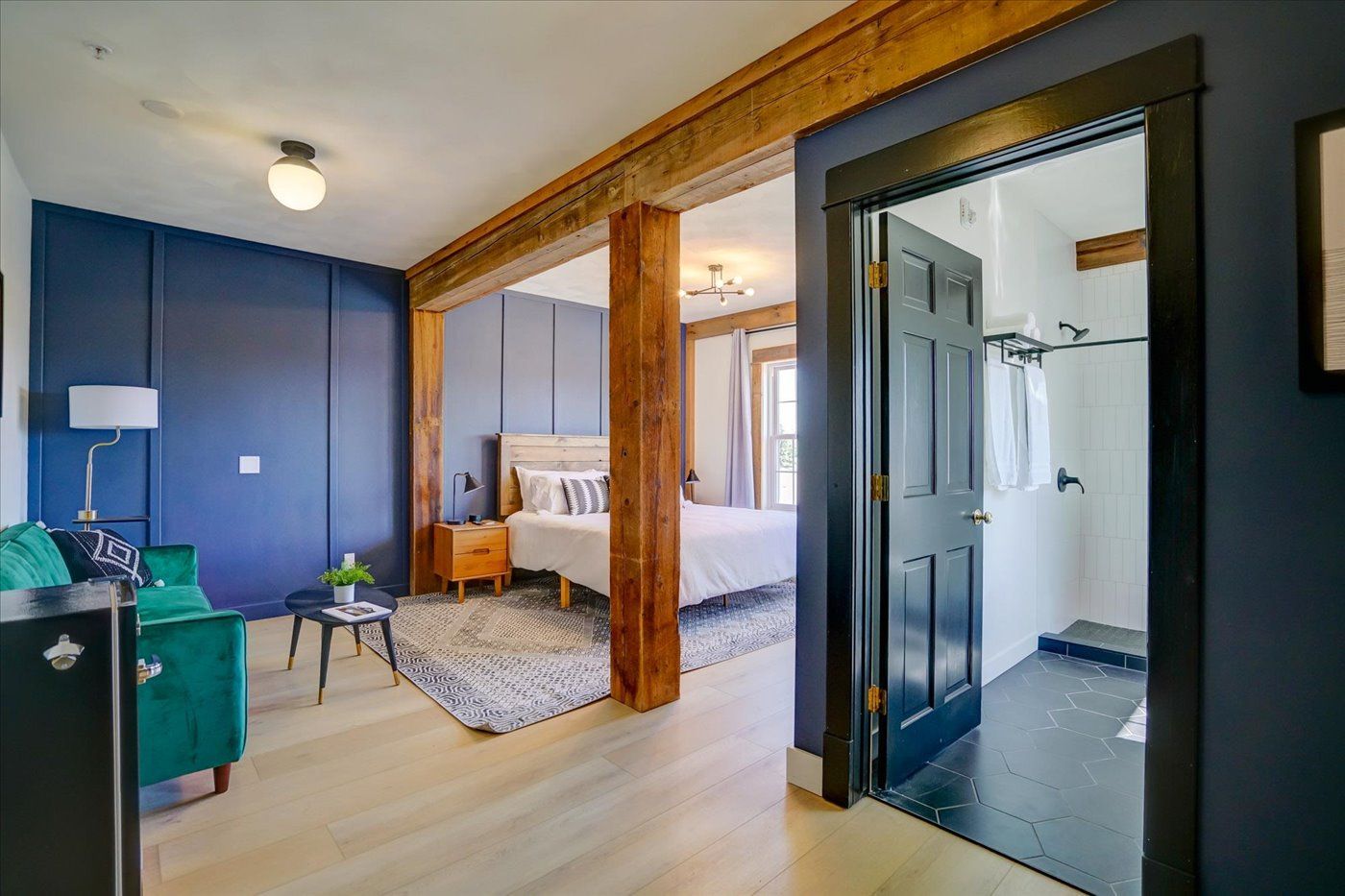An open-plan hotel suite with blue walls, a bedroom, and a bathroom. Includes wooden beams and light wood floors.