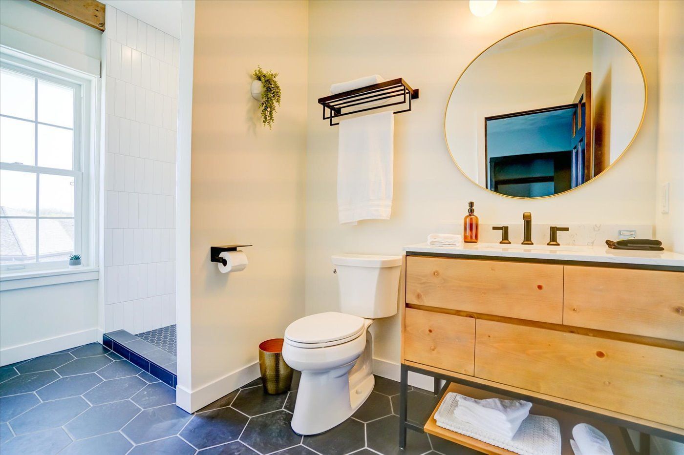 Bathroom with a wooden vanity, round mirror, white toilet, and dark hexagon tile floor.