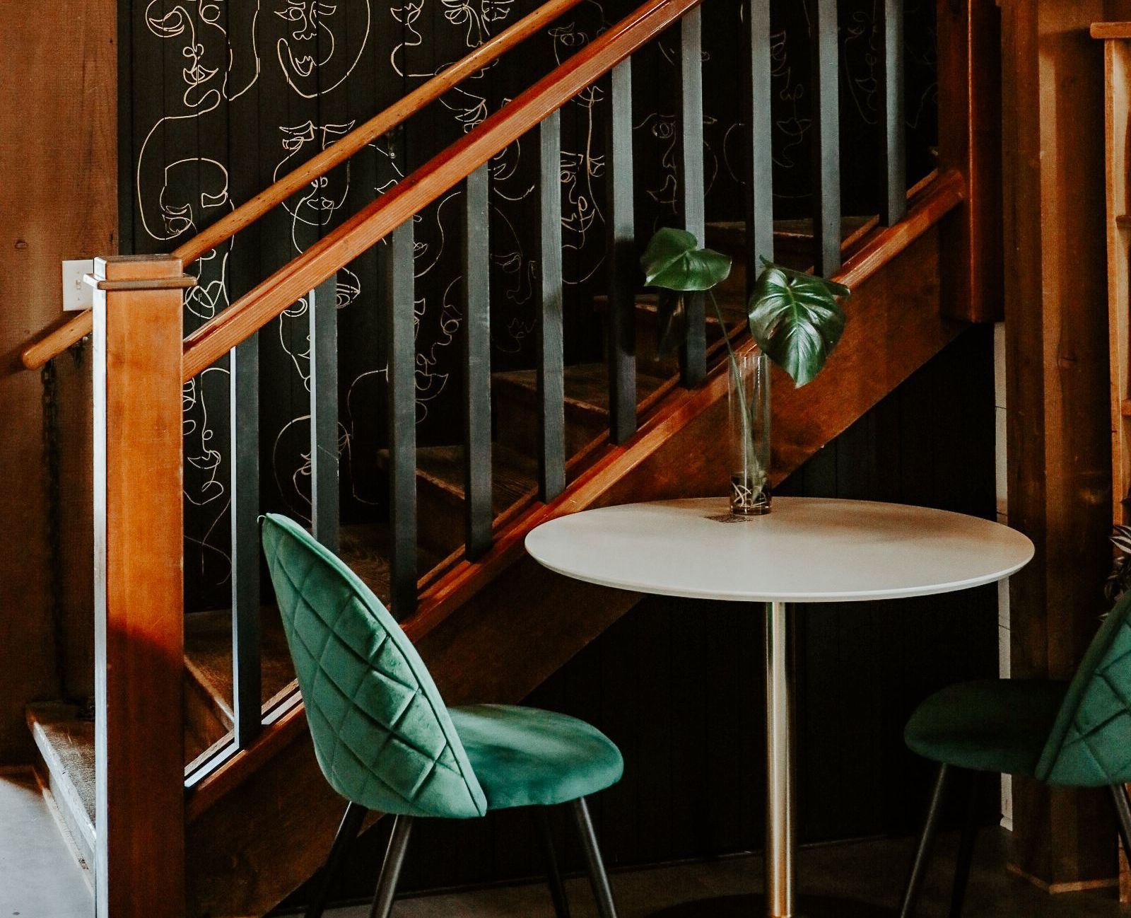 Two green chairs at a round table, under wooden stairs with black railings.