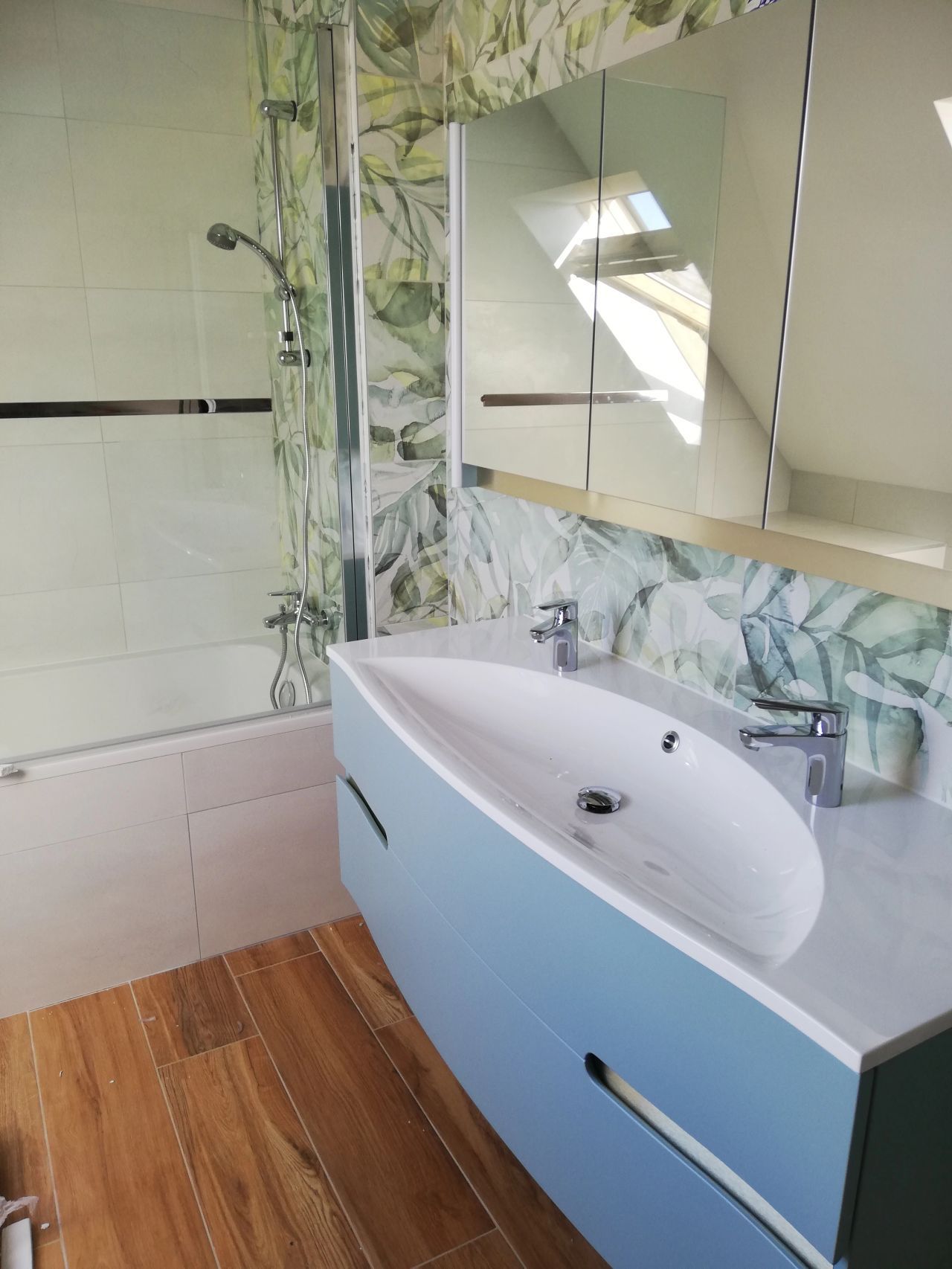 Salle de bains moderne avec vanité bleu clair, lavabo blanc, douche en verre et plancher à imitation en bois.