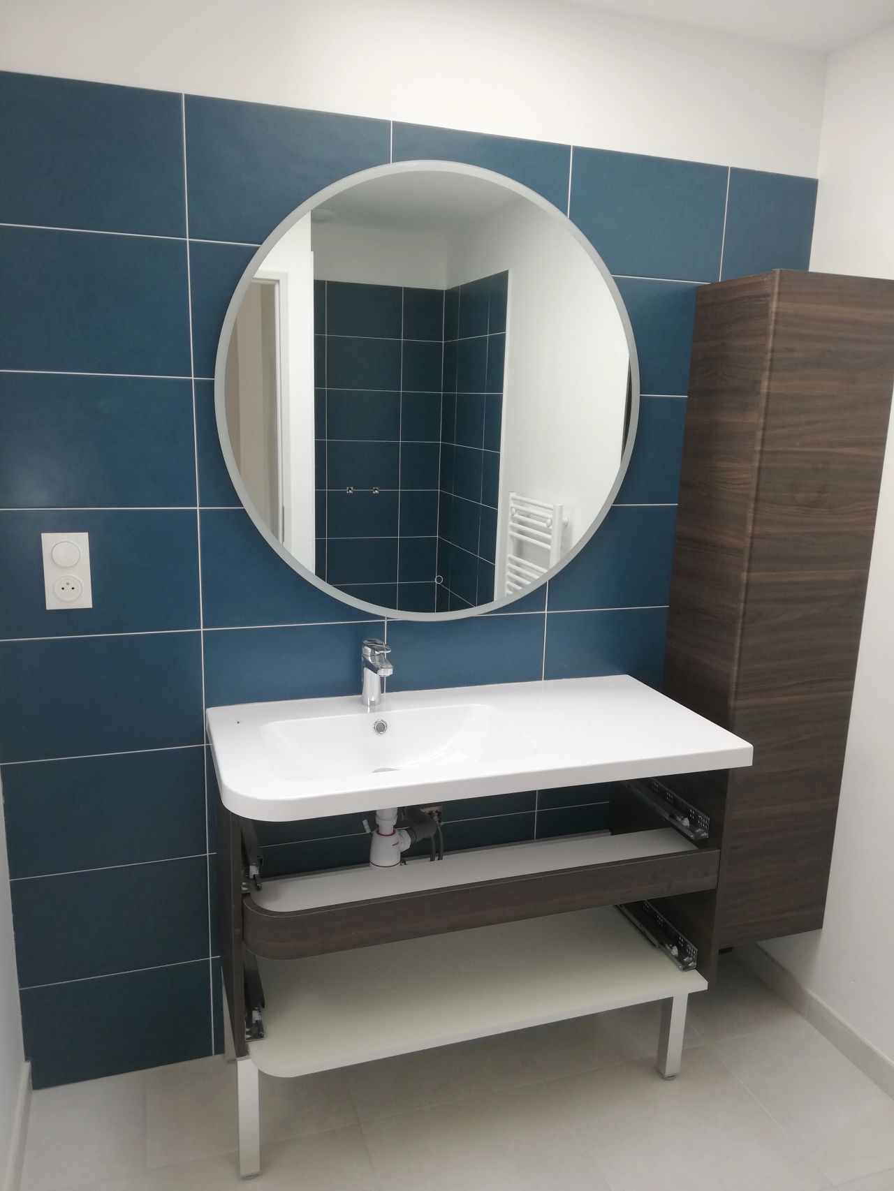 Salle de bains avec mur carrelé bleu, miroir rond, lavabo blanc et armoire marron.
