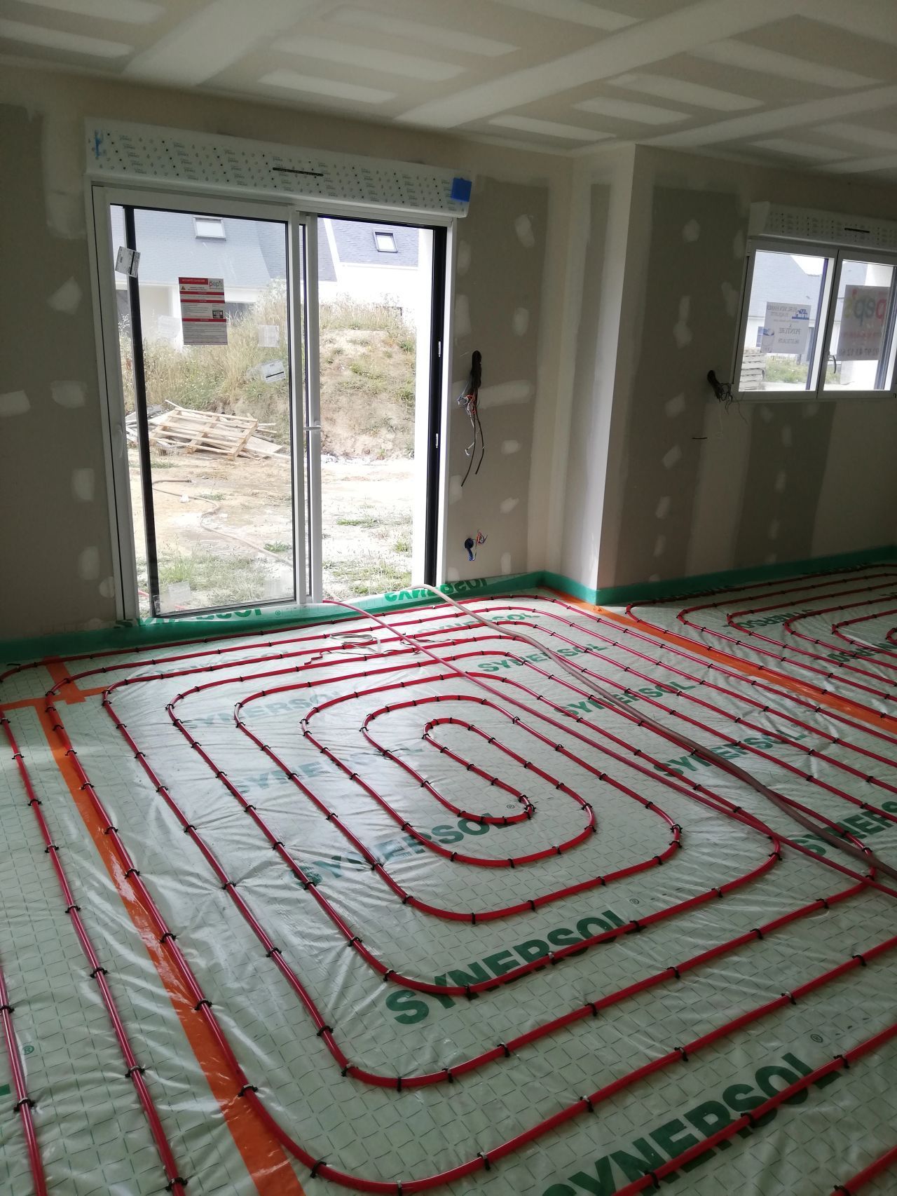 Une pièce en construction ; des tubes rouges pour le chauffage par le sol sont installés sur une feuille de plastique verte.