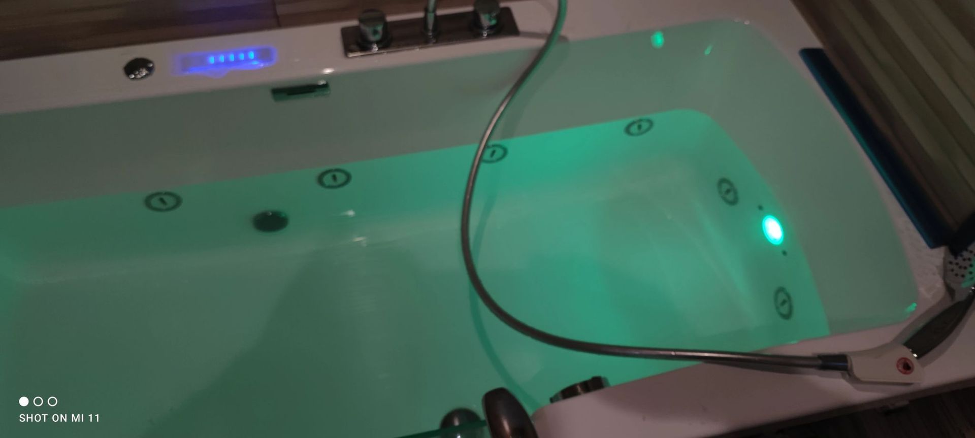 Baignoire blanche remplie d'eau éclairée en vert. On aperçoit une douchette et un robinet argentés.