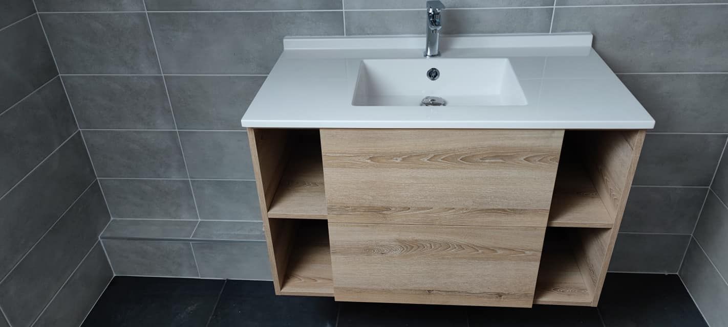 Lavabo de salle de bain avec comptoir blanc, armoire en bois clair avec étagères ouvertes et murs en carrelage gris.