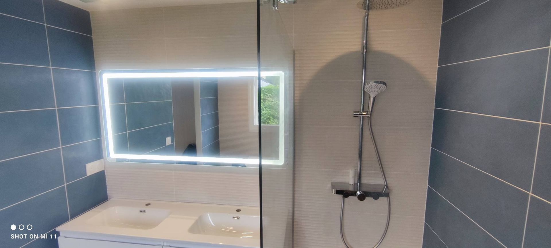 Salle de bains avec carrelage bleu, porte de douche en verre et miroir avec bordure lumineuse au-dessus d'un lavabo blanc.