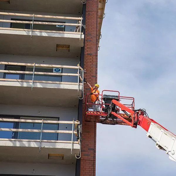 Oranssiin varusteisiin pukeutunut työntekijä kirsikanpoimijalla korjaa tiilirakennuksen parveketta