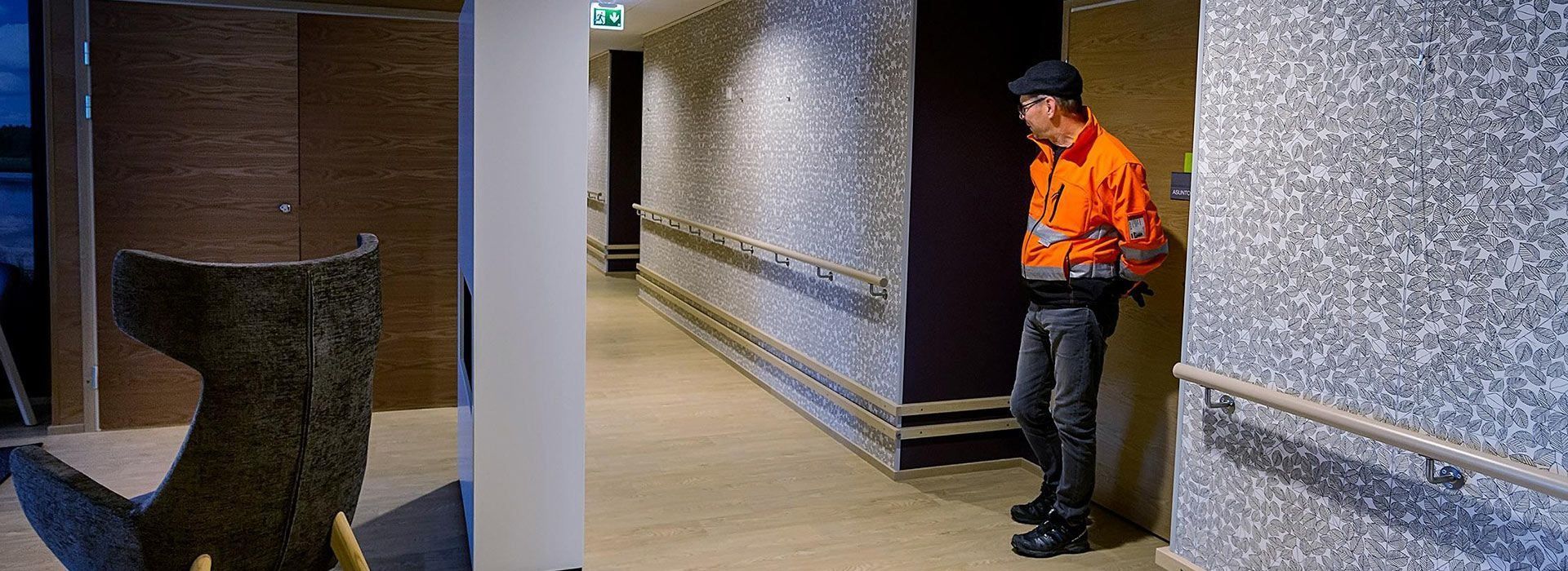 Oranssiin takkiin pukeutunut henkilö seisoo laatoitetulla käytävällä seinän vieressä.