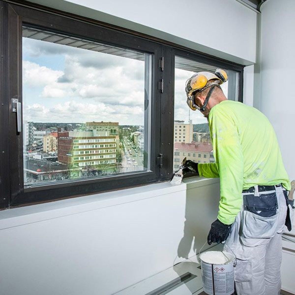 Keltaisiin turvavarusteisiin pukeutunut työntekijä maalaa valkoista seinää ikkunan vieressä, josta on kaupunkinäkymä
