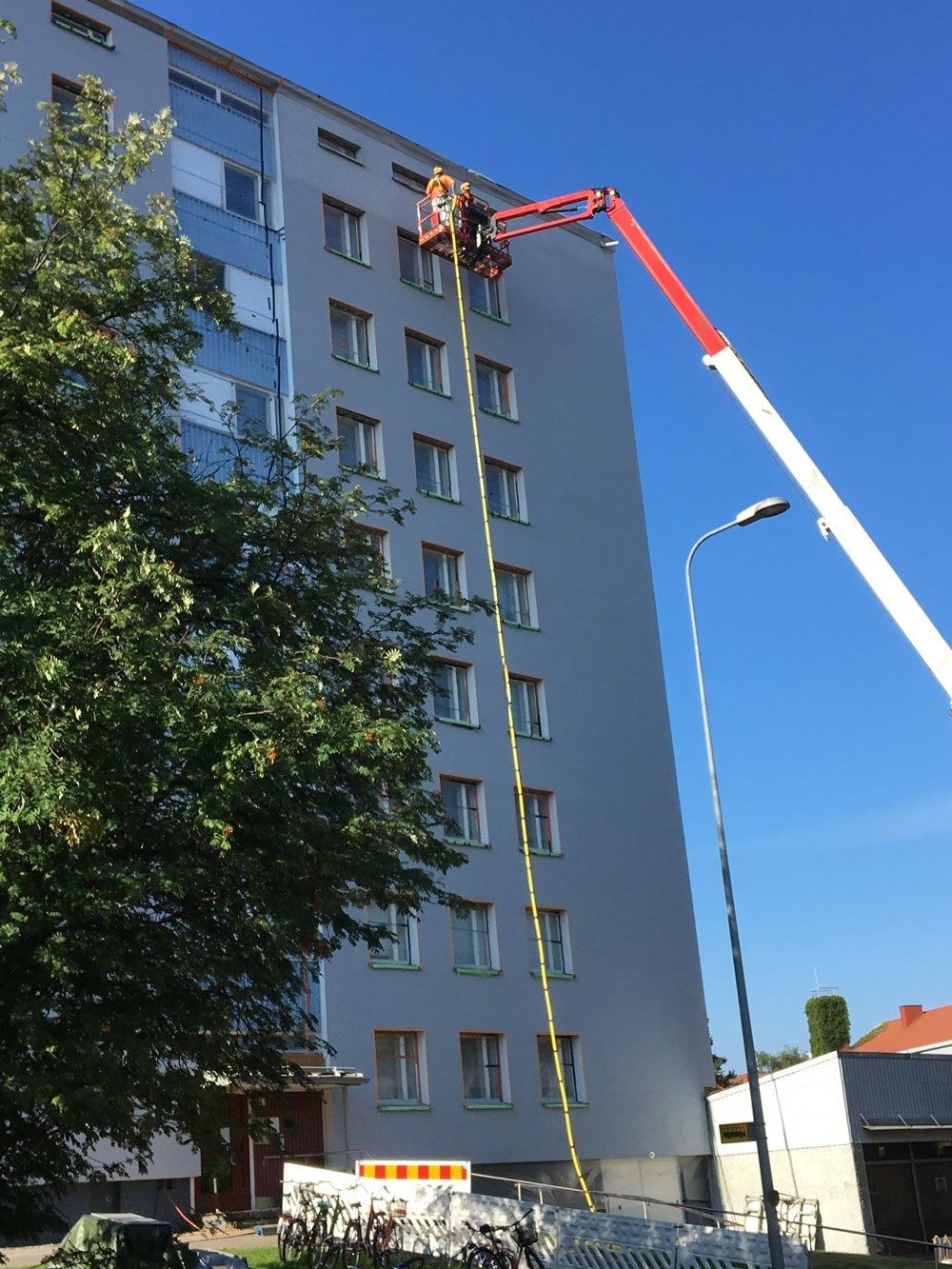 Punaisessa puominostimessa oleva työntekijä korjaa harmaan kerrostalon yläkerroksia kirkkaana päivänä.