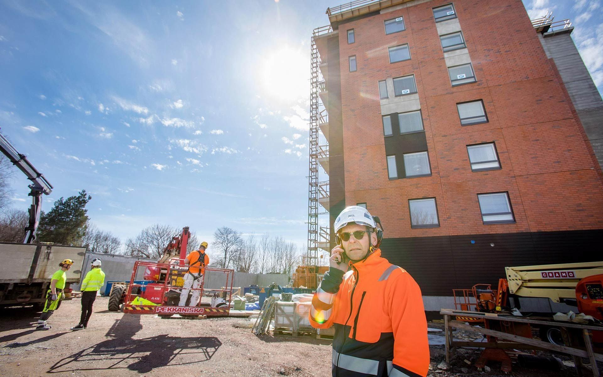 Rakennustyöläisiä oranssiin turvavarusteisiin pukeutuneina korkean tiilirakennuksen vieressä, nosturin ja kirkkaan taivaan edessä.