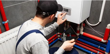 Un técnico con gorra ajusta el panel de control de una caldera mientras sostiene una tableta. Se ven tuberías rojas y azules.