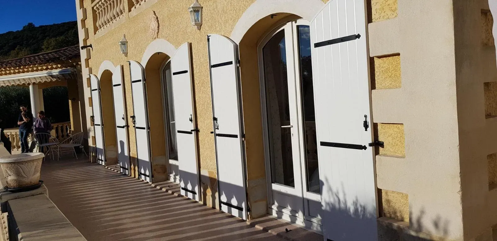 Par une journée ensoleillée, on aperçoit un bâtiment jaune aux fenêtres cintrées, aux volets blancs et à la terrasse en pierre.