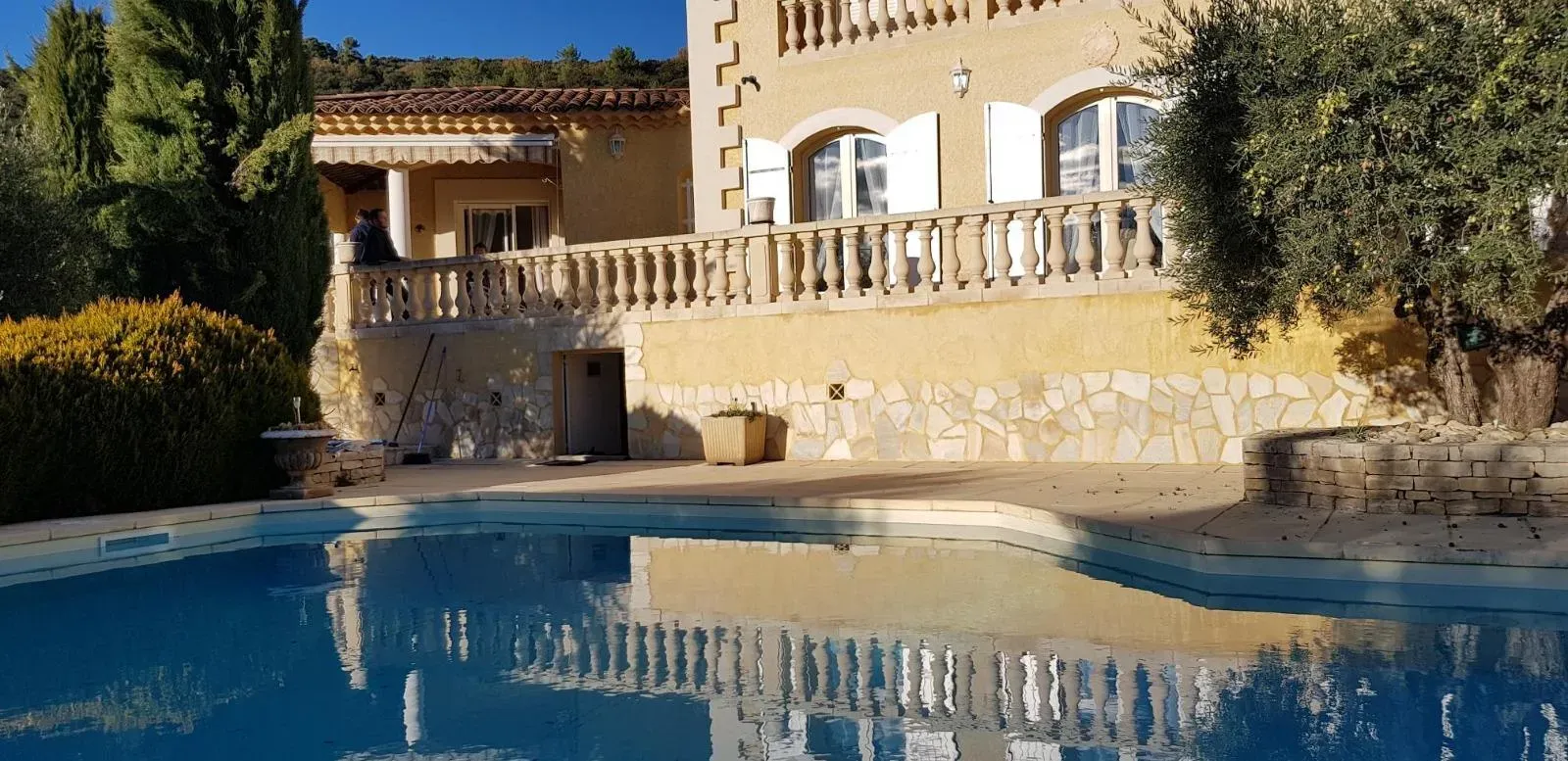 Piscine devant un bâtiment en pierre aux volets blancs et doté d'un balcon. Un arbre se trouve à droite.