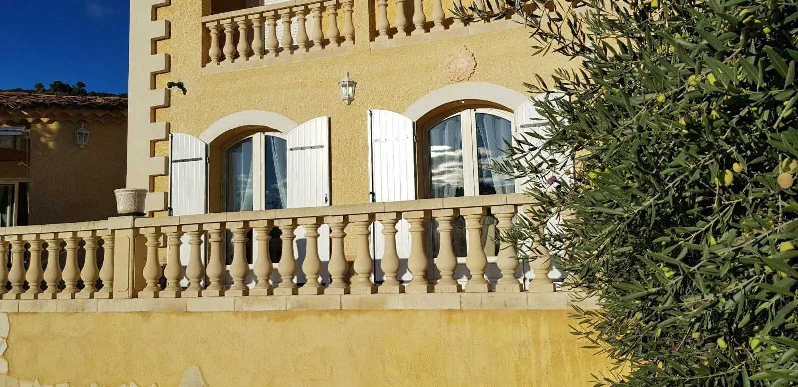 Bâtiment en stuc jaune avec fenêtres cintrées, volets blancs et balustrade en pierre. Un olivier se trouve sur le côté droit.
