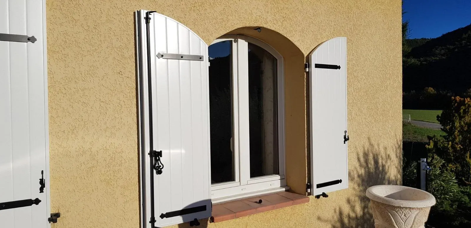 Volets blancs encadrant une fenêtre à arc surbaissé, sur un mur en stuc jaune.