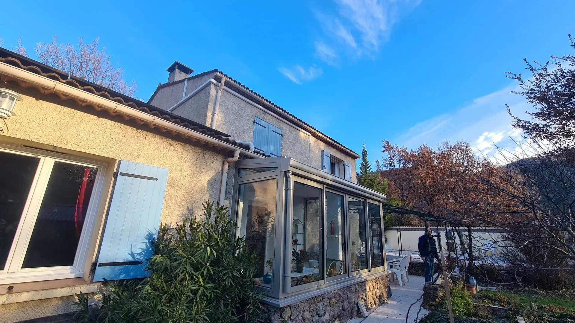 Extérieur de la maison avec véranda vitrée, volets bleus et ciel dégagé