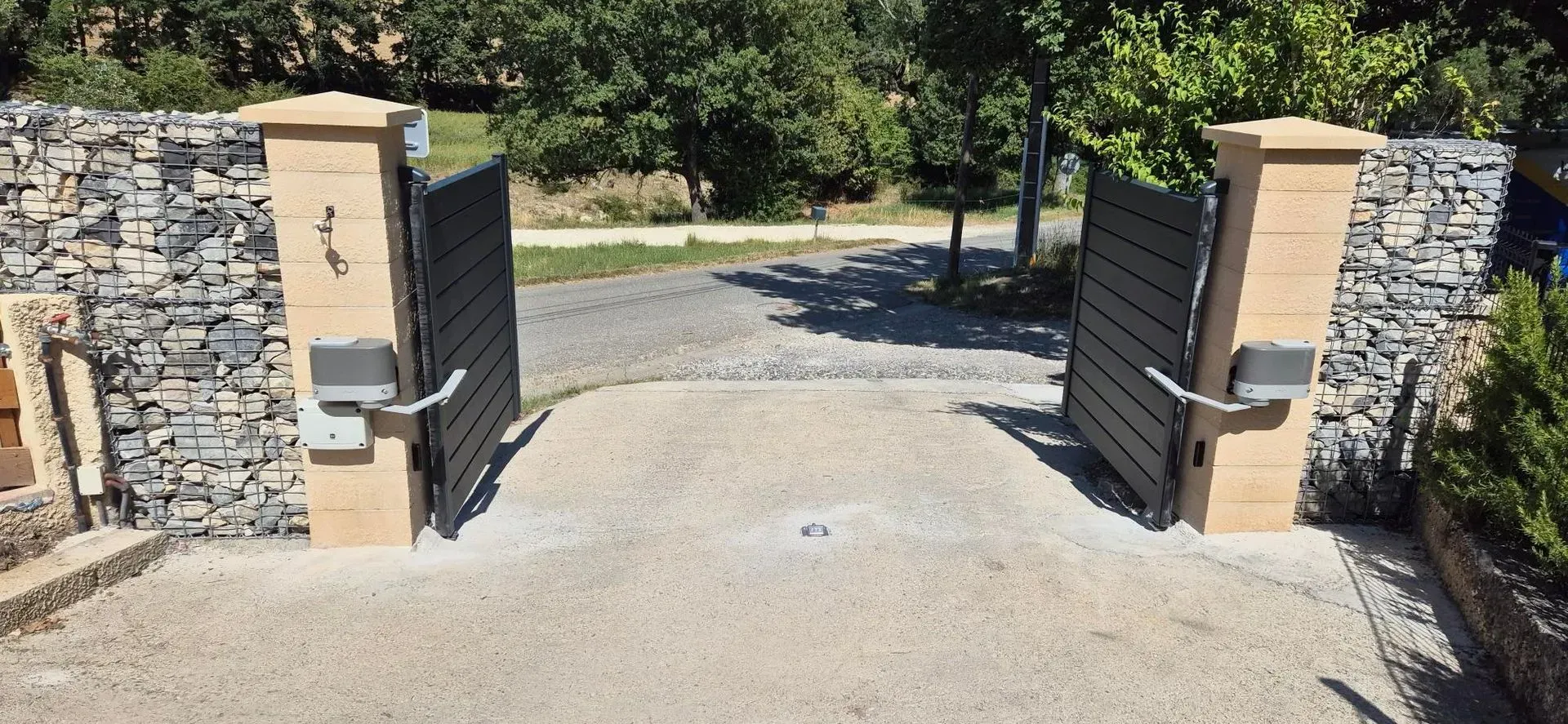 Des portails gris foncé ouverts entre des piliers de pierre donnent sur une allée de gravier.