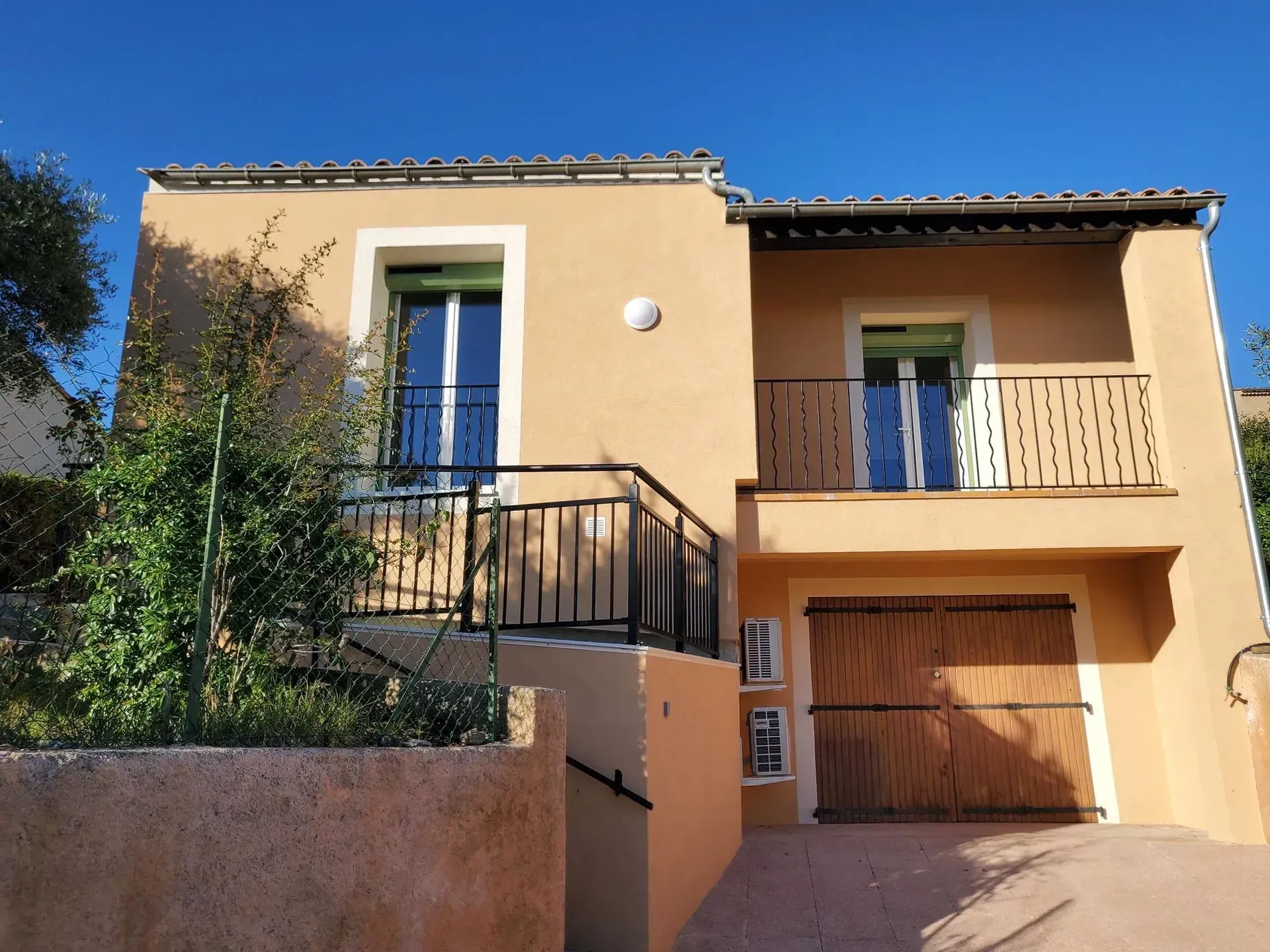 Maison beige avec portes bleues, balustrades noires et allée carrossable, sous un ciel bleu azur.