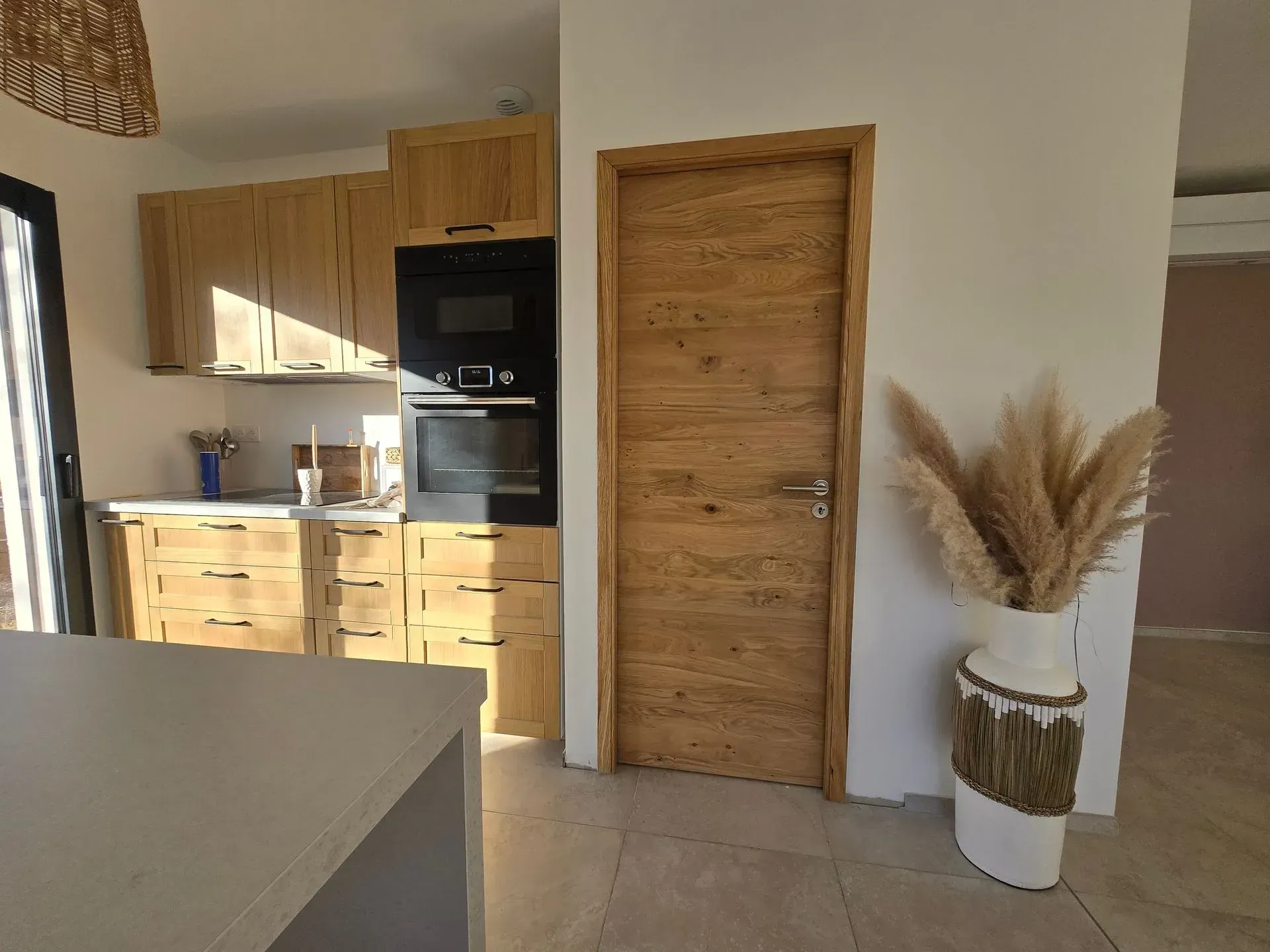 Cuisine avec placards en bois clair, four et porte en bois. Un vase contenant de hautes herbes séchées se trouve à proximité.