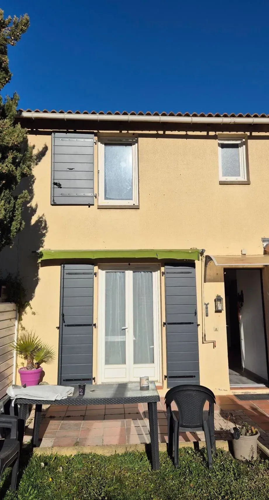 Bâtiment jaune de deux étages avec des volets gris et un ciel bleu ; table et chaises à l'extérieur, devant.
