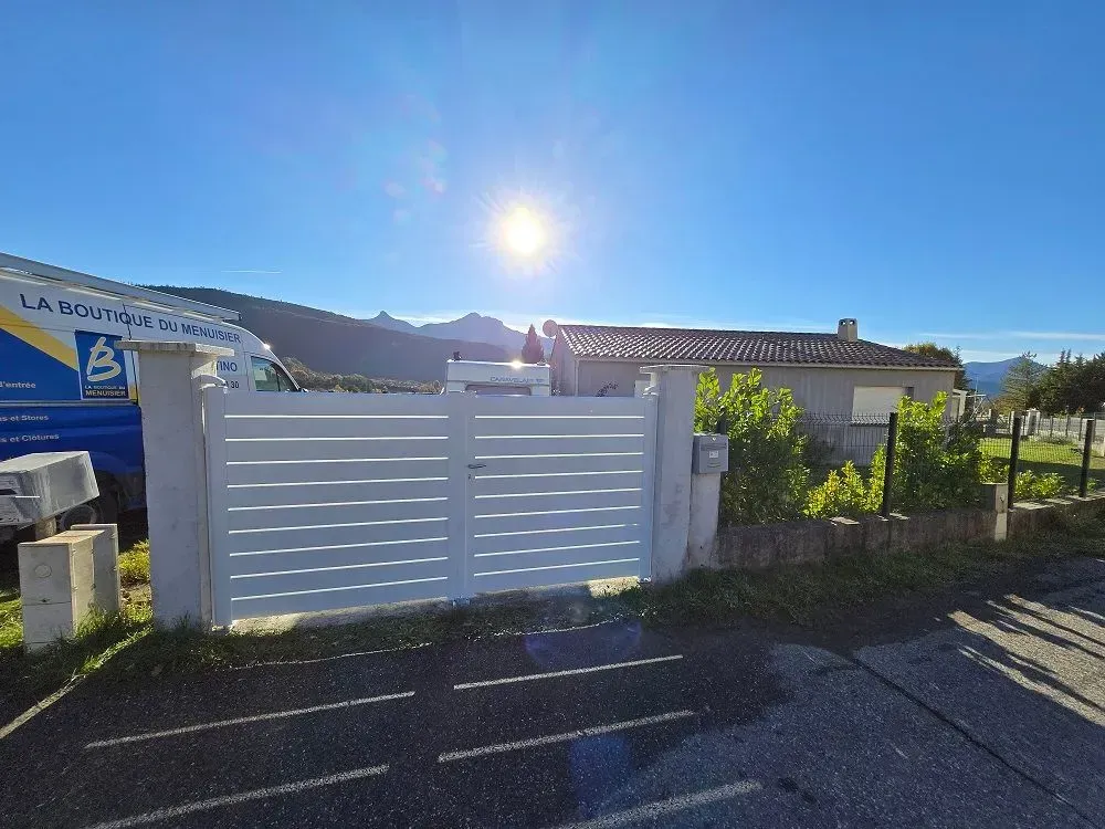 Portail à lattes blanches avec des poteaux en béton devant une maison, soleil éclatant dans le ciel.