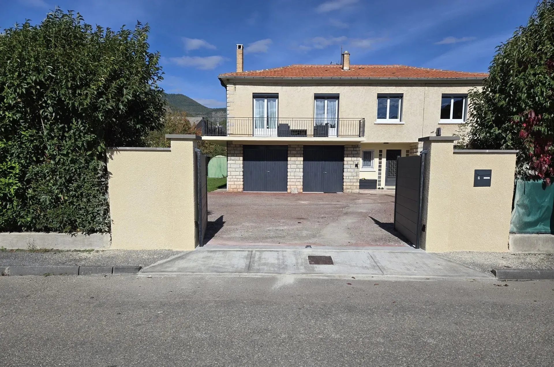 Maison beige à deux étages avec une allée privée dissimulée derrière un mur. Deux portes de garage sont visibles. Des arbres bordent la maison.
