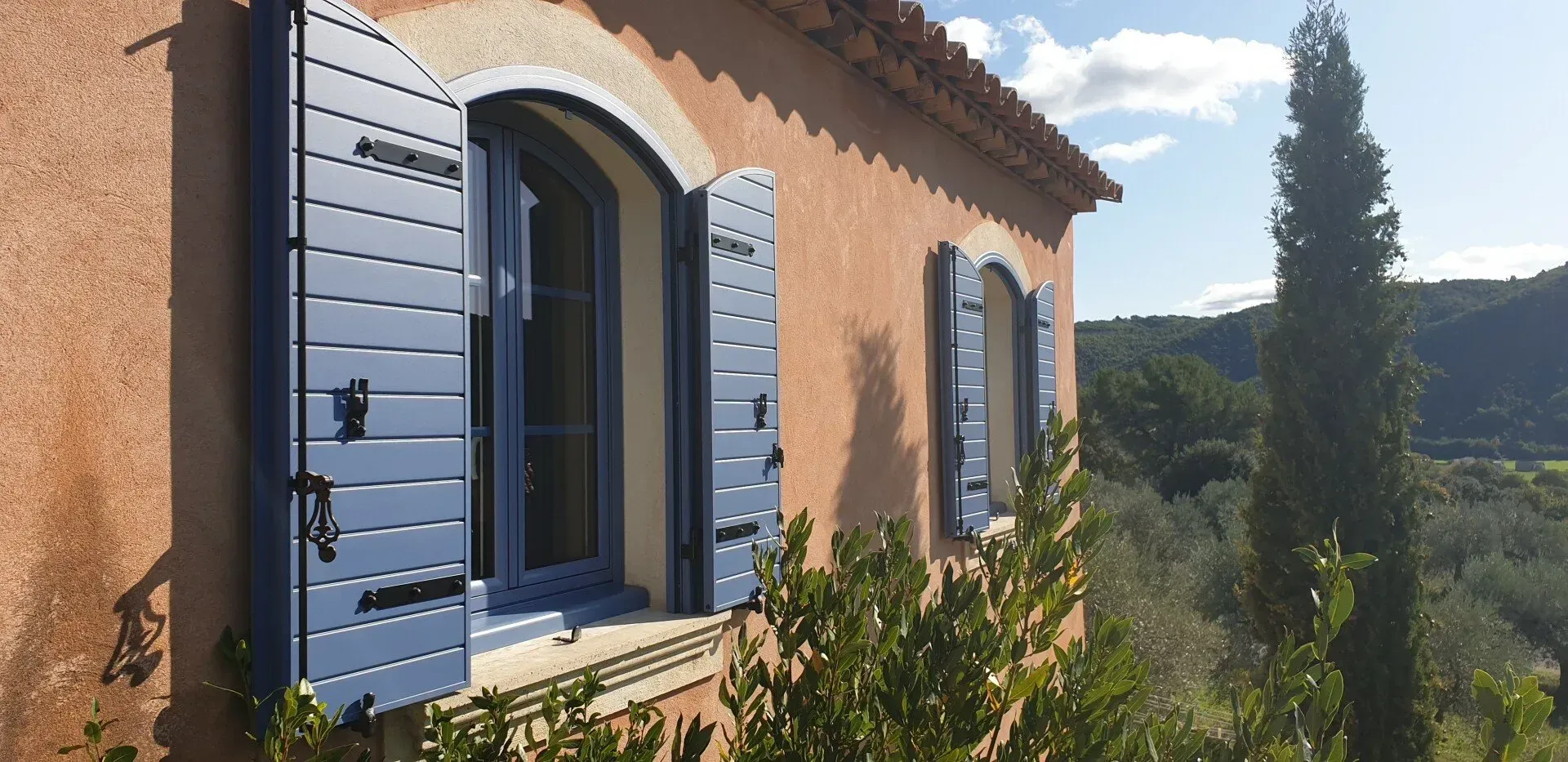 Un bâtiment aux volets et fenêtres bleus, entouré d'arbres et d'une colline.