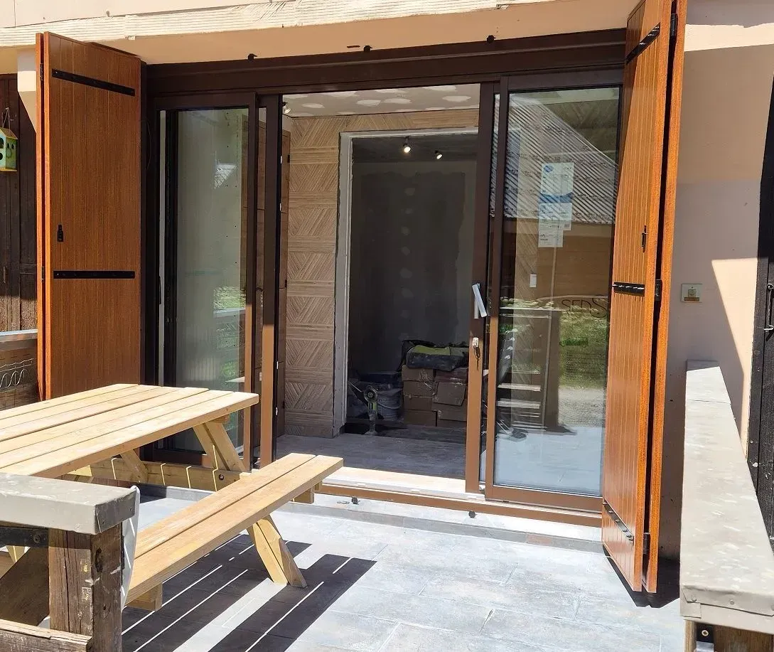 Vue extérieure : Porte marron ouverte avec panneaux de verre ; table de pique-nique en bois.