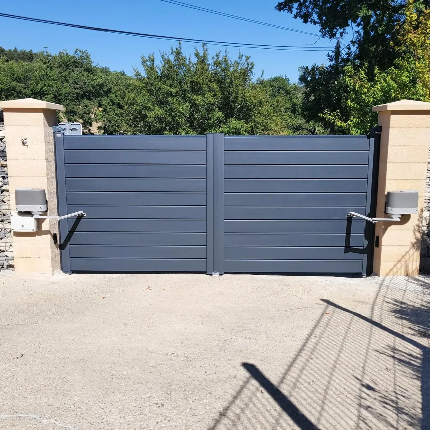 Portail double gris motorisé sur piliers en pierre, allée de gravier et feuillage en arrière-plan.