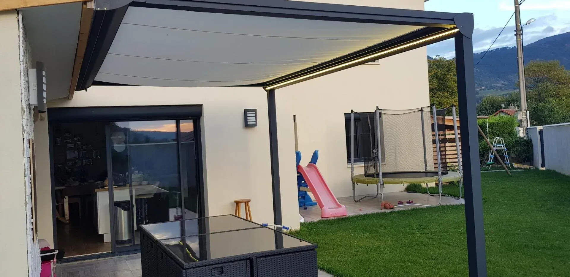 Une terrasse moderne avec un auvent rétractable, une maison, un jardin avec des jeux et des montagnes en arrière-plan.