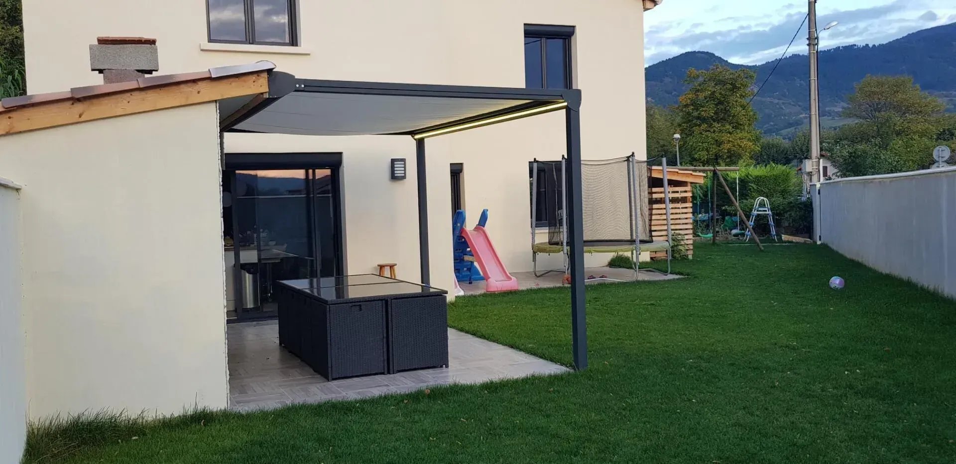 Une maison moderne avec une pergola, du mobilier de jardin noir et un jardin gazonné.
