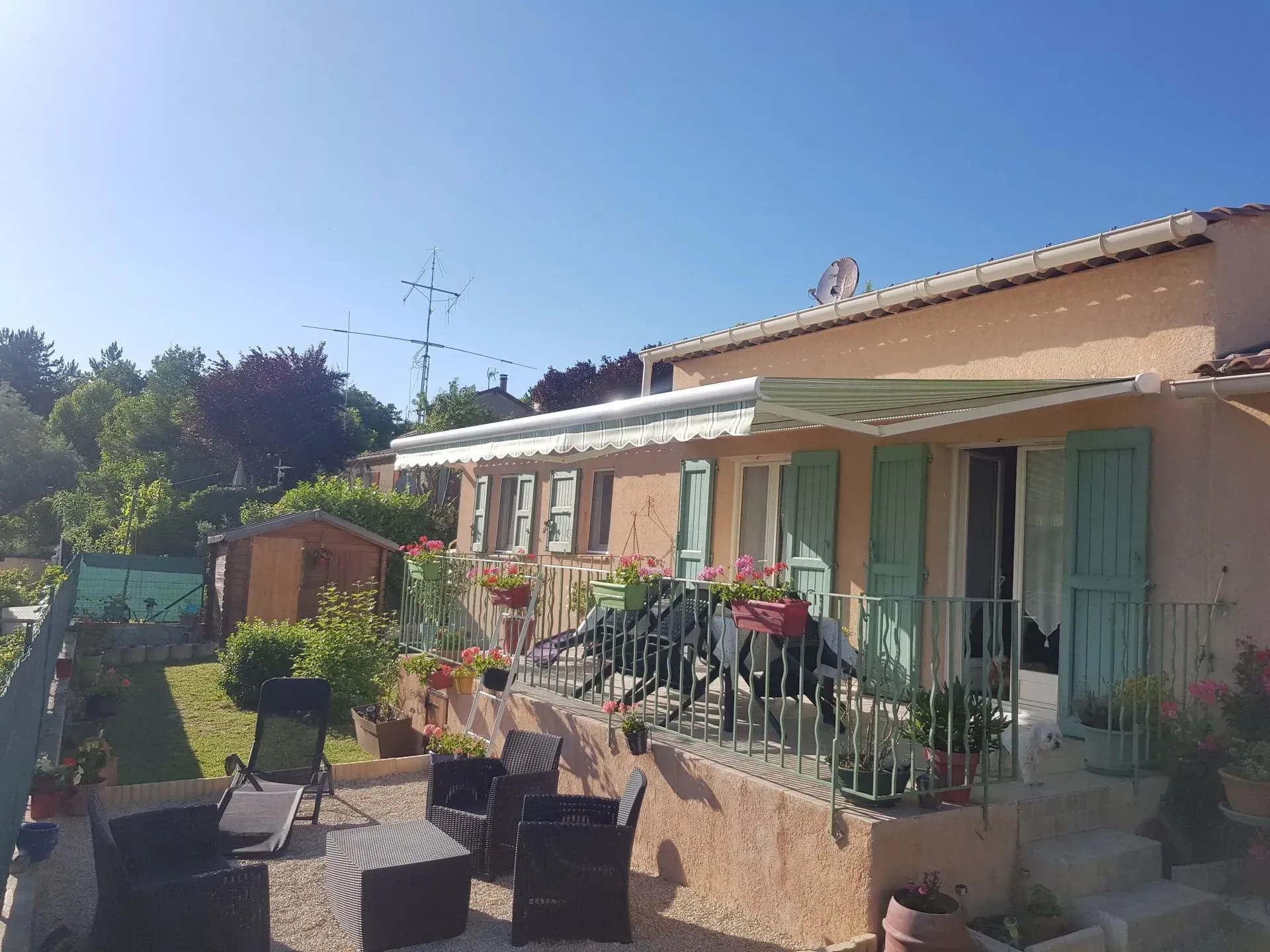 Maison à la façade beige clair, volets verts, auvent et terrasse avec chaises sous un ciel bleu azur.
