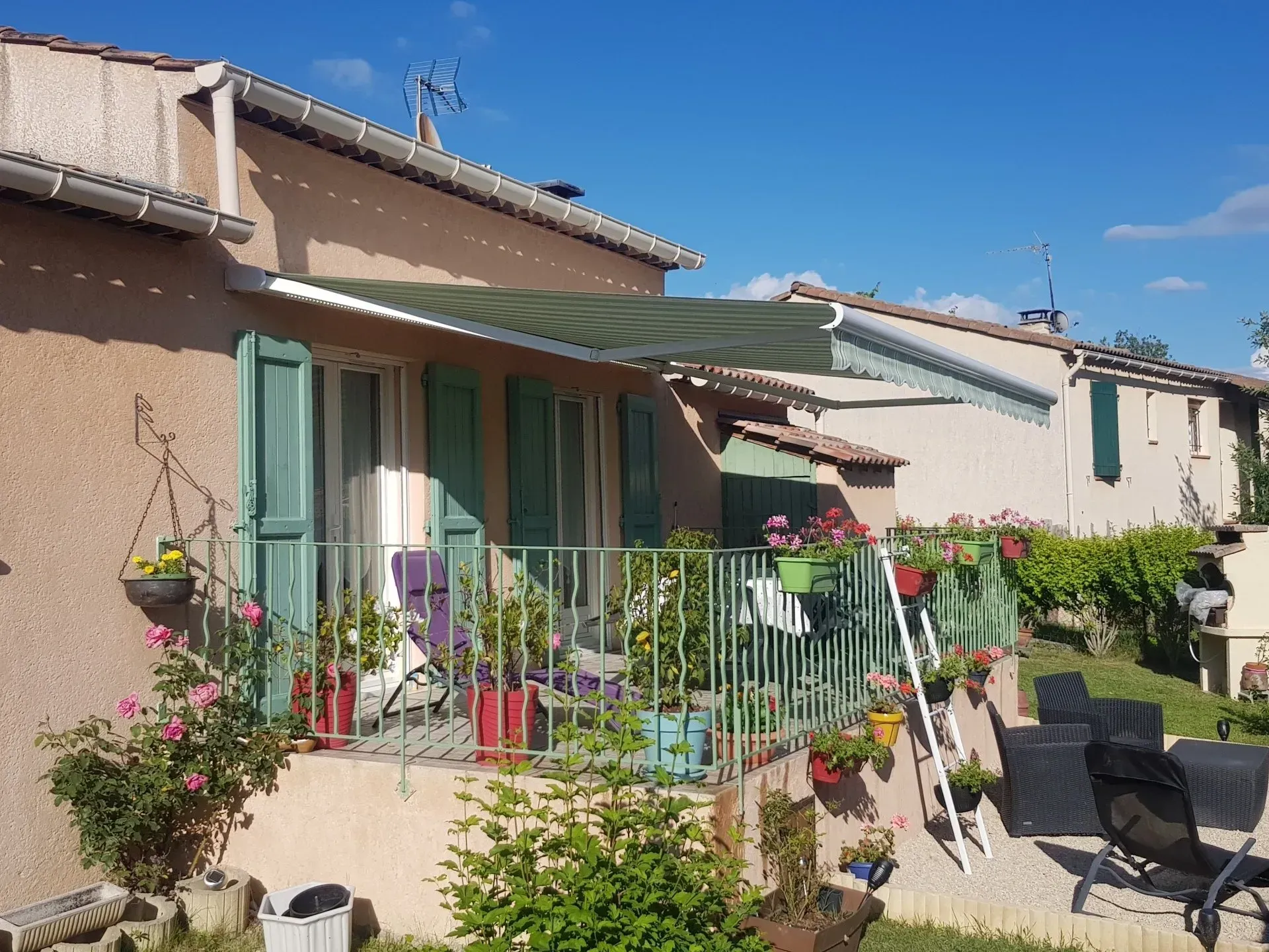 Immeuble beige avec balcon, auvent vert, volets et plantes fleuries. Ciel bleu.